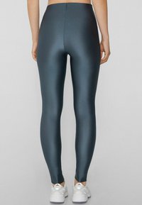 Leggings taille haute, en teal foncé, avec une texture lisse et brillante. Coupe ajustée et finition sans couture, associés à des baskets blanches.