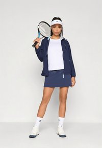 Navy tennis outfit featuring a zippered jacket, white shirt, and a matching skirt, complemented by a tennis racket and white athletic shoes.