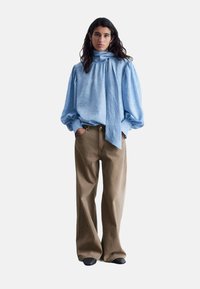 Helles blaues, strukturiertes Blusenoberteil mit einem gebundenen Kragen, kombiniert mit beigen, weit geschnittenen Hosen und schwarzem Schuhwerk. Minimalistisches Design, lässige Passform.
