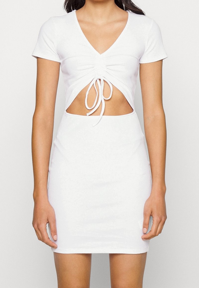 Femme portant une robe blanche à manches courtes avec un profond décolleté en V, un devant plissé et une découpe au niveau du ventre, debout devant un fond uni.