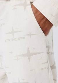 Main dans la poche d'un pantalon clair avec des motifs subtils d'étoiles et un design de texte "PXP" répété.