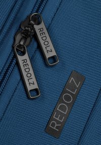 Blauwe stoffen ritszak met twee zwarte ritsen waarop "REDOLZ" is gegraveerd, en een zwart leren label met het reliëflogo "REDOLZ".