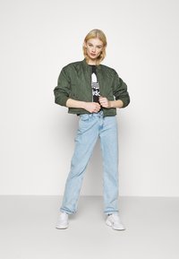 Kvaliteediga oliivroheline bomber-jakk, millel on soonikkoes varrukad, kantud musta graafilise t-särgi üle, koos helesiniste sirgete pükstega ja valgete jalgadega.