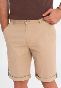 Shorts en coton beige avec poches latérales, fermeture à bouton et ourlets retroussés rehaussés d'un détail bleu. Texture lisse, longueur au milieu de la cuisse.
