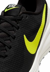 Mustat urheilukengät, joissa on teksturoitu päällinen, kirkas keltainen Nike-logomerkki, mustat nauhat ja valkoinen, kaareva välipohja.