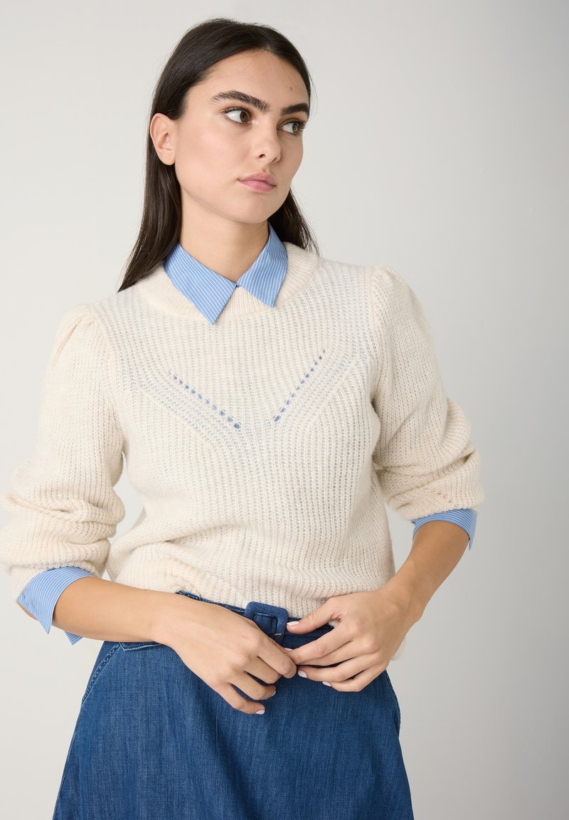Cremfarbener Strickpullover mit gerippter Textur und blau gestreifter Kragenbluse darunter. Modell trägt einen Jeansrock und passt die Gürtelschließe an.