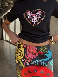 Μαύρη μπλούζα με σχέδιο λουλουδιών σε σχήμα καρδιάς σε ροζ και πράσινο. Πολύχρωμη φούστα με σχέδιο που περιλαμβάνει διάφορα φυτά και λουλούδια.