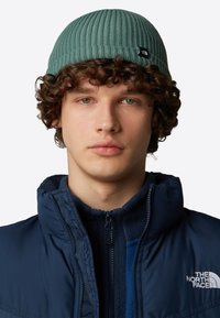 Beanie a coste verde realizzato in materiale morbido, con un piccolo cartellino con logo nero. Indossato con una giacca blu navy con zip sopra uno strato in pile.