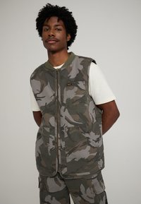 Chaleco de camuflaje con cierre delantero de cremallera, cuello de canalé verde, múltiples bolsillos frontales y un ajuste holgado. Fabricado con tela resistente.