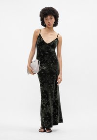 Zelený zamatový šaty s kvetinovým vzorom, výstrih do V, tenké ramienka, bočný rozparok, doplnené svetlošedou kabelkou a čiernymi sandálmi.