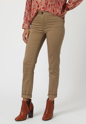 Pantalones ajustados color beis con cintura media, con puños enrollados. Llevados con botas de tobillo marrones, destacando una textura de tela suave.