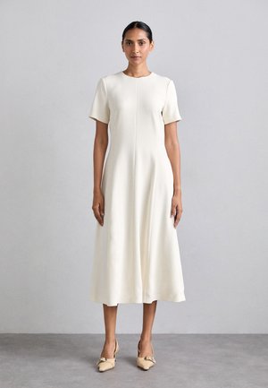 Vestido creme de mangas curtas com decote redondo, comprimento até à meia-panela, e uma textura suave. Combinado com sapatos beiges tipo slip-on com um detalhe de laço.