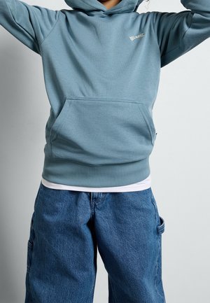 Personne portant un sweat à capuche Billabong bleu clair et un jean bleu baggy, les mains levées, cadrée au-dessus de la bouche et en dessous de la taille.
