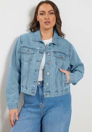 Frau mit langen braunen Haaren, die eine hellblaue Jeansjacke über einem weißen Hemd und blauen Jeans trägt, steht vor einer weißen Wand.