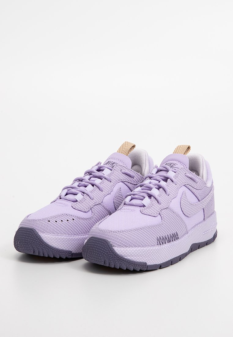 Zapatillas de deporte de color púrpura claro con parte superior de malla texturizada, suela de goma y lengüeta de color marrón. Presenta un diseño de cordones y el logo de Nike en el lateral.