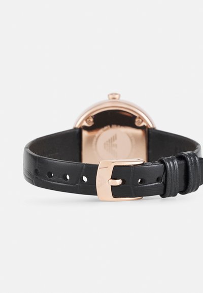 Montre ronde avec un boîtier en or rose et un bracelet en cuir noir. Présente un bracelet texturé, une boucle en métal et un fond lisse et poli.