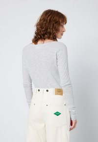 Camisola cinza de mangas longas combinada com jeans de cintura alta em off-white, apresentando um patch em forma de diamante verde e uma etiqueta de couro castanho na parte de trás.