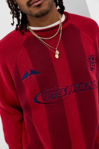 Camisola vermelha de malha com gola canelada, com logótipo azul bordado e gráfico no peito, combinada com colares dourados em camadas.