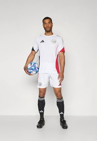White soccer jersey with red accents, black shorts, and black socks, featuring an emblem and three-stripe branding. Holds a multicolored soccer ball.