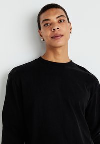 Camiseta negra de manga larga hecha de tela de algodón con cuello redondo. Presenta un ajuste holgado y un diseño minimalista. No tiene patrones ni acentos visibles.