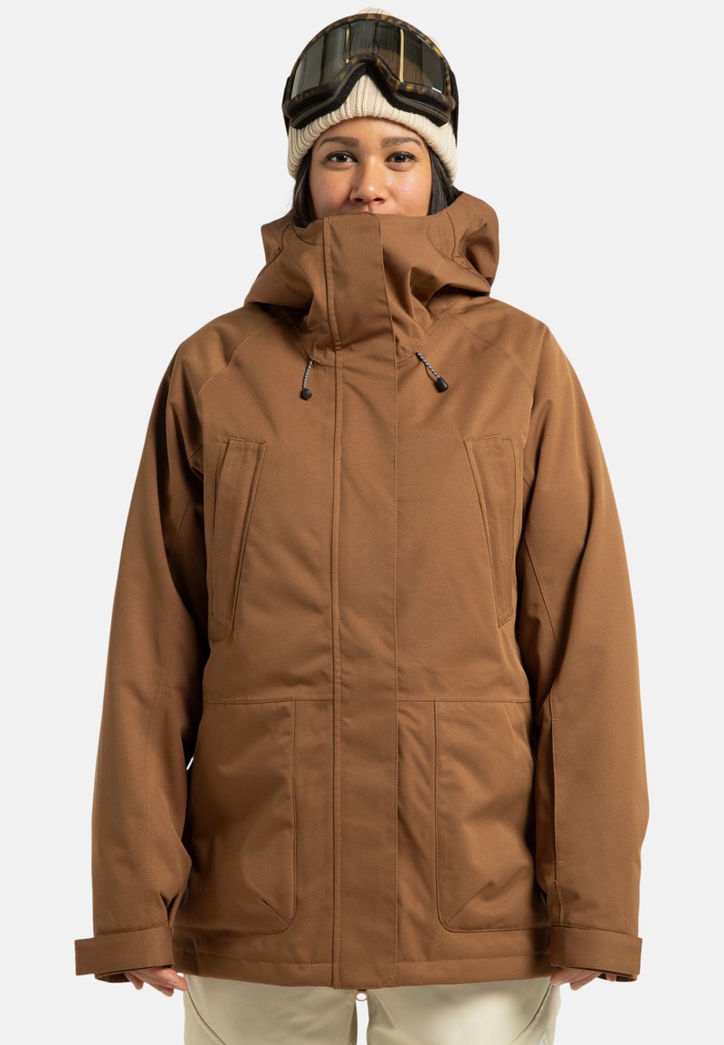 Braune wasserdichte Jacke mit hohem Kragen, mehreren Taschen und verstellbaren Kordelzügen. Getragen mit gestrickter Mütze und Skibrille.