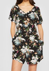 Mujer vistiendo un mono corto de manga corta y escote en V negro con cinturón y bolsillos, que presenta patrones de flores tropicales en blanco, verde, rosa y azul.