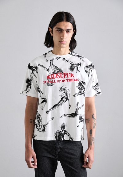 T-shirt en coton blanc avec des figures dessinées au crayon noir et le texte rouge "KIDSUPER IT'S ALL UP IN THE AIR." Présente une coupe décontractée et un col rond.