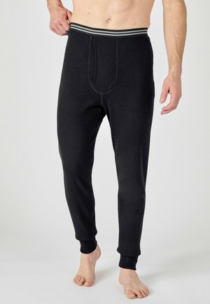 Mann trägt schwarze lange Unterhose mit gestreiftem elastischem Bund, steht barfuß vor hellem einfarbigem Hintergrund.
