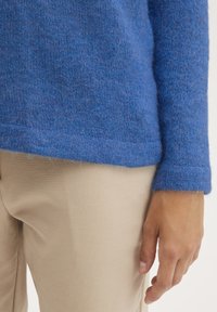 Zils adīts džemperis ar mīkstu, pūkainu tekstūru un nedaudz brīvu piegriezumu, apvienots ar bēšām biksēm, izceļot piedurkņu un apakšmalu detaļas.