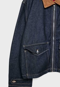 Veste en denim avec un col en velours côtelé marron, deux poches avant avec des fermetures à pression et des coutures contrastantes visibles. Coupe courte et carrée.