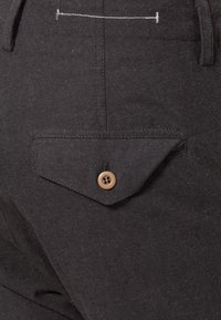 Gros plan sur un pantalon en tissu foncé montrant une poche à rabat boutonnée et des passants de ceinture visibles avec une couture horizontale blanche au-dessus de la poche.