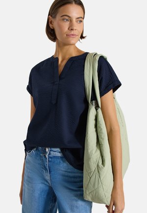 Kobieta w granatowej, teksturowanej bluzce z krótkim rękawem i niebieskich jeansach, niosąca na ramieniu dużą, pikowaną, jasnografitową torbę.