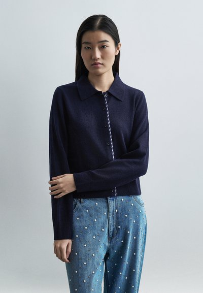 Jeune femme aux longs cheveux foncés portant un cardigan bleu marine boutonné et un jean bleu orné de perles blanches.