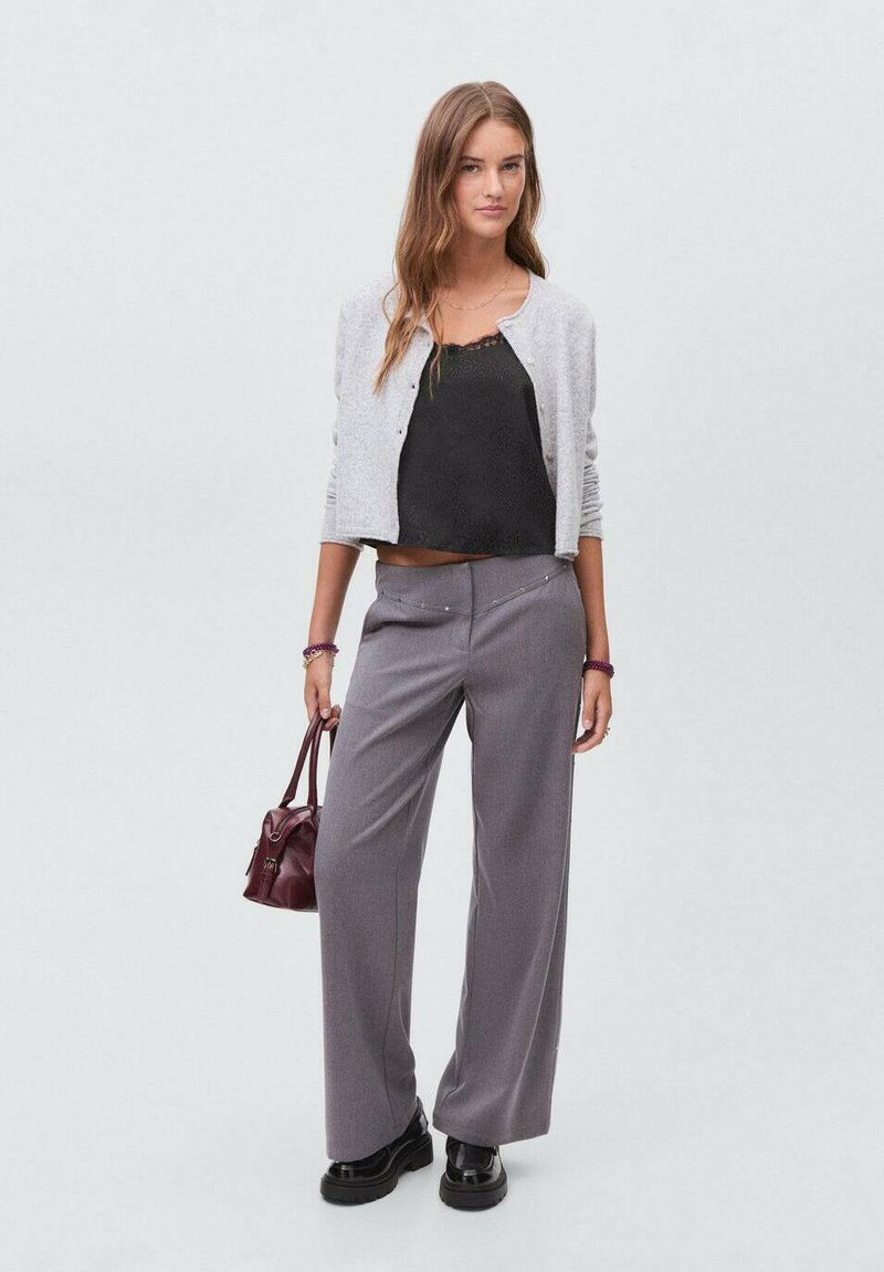 Siva pletena jopica, črna top z naramnicami in ohlapne sivo hlače z dekorativnimi špiki. Model drži bordo ročno torbico in nosi masivne črne čevlje.