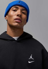 Felpa nera con loghi bianchi di Jordan e Nike, berretto azzurro a coste con texture e orecchini a cerchio argentati. Veduta ravvicinata della parte superiore del corpo e del viso.