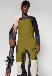 Macacão de esqui em verde oliva com detalhes em preto e creme, apresentando bolsos com fecho e alças ajustáveis. O modelo segura um esqui com ligações.