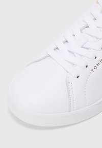 Fehér sneaker fehér fűzővel, sima orr résszel és diszkrét varrási részletekkel az oldalon, részben látható márkás szöveggel.