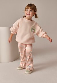 Salopetă din bumbac roz cu un design de desene animate, asortată cu colanți evazați în dungi portocalii și albe. Pantofii de culoare deschisă completează ținuta.