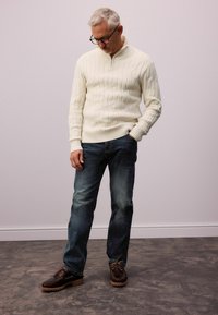 Suéter de punto de cable color crema con cremallera de cuarto, combinado con jeans azul oscuro y zapatos marrones con suela texturizada y cordones.