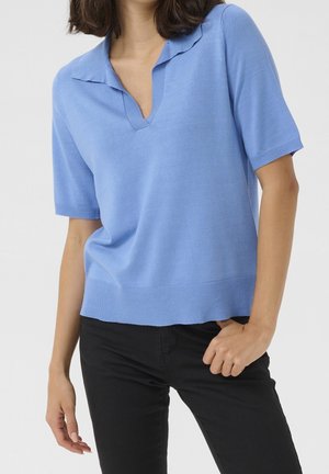 Woman wearing a light blue short-sleeve knit top with a collar and deep V-neck, paired with black pants against a neutral background.