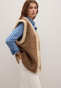 Braune Kunstwildlederweste mit einem cremefarbenen Schaffellfutter. Bietet einen lässigen Schnitt, offenen Front und Seiten Taschen, kombiniert mit einem blau gestreiften Hemd.