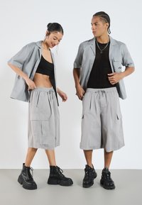 Deux mannequins portent des chemises à manches courtes grises assorties sur des tops noirs, associées à des shorts cargo gris clair et des bottes à plateforme noires.
