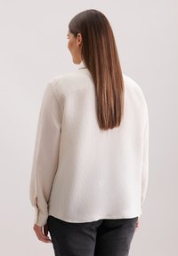 Blusa a coste color crema con maniche lunghe. Presenta polsini con bottoni e orlo arrotondato. Indossata sopra pantaloni scuri.