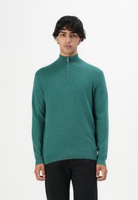 Junger Mann, der nach vorne blickt und einen türkisfarbenen Pullover mit halbem Reißverschluss sowie schwarze Hosen trägt, vor einem schlichten weißen Hintergrund.