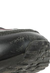 Gros plan d'une semelle de chaussure noire usée avec saleté visible, éraflures et fibres effilochées sur la bande de roulement sur fond blanc.