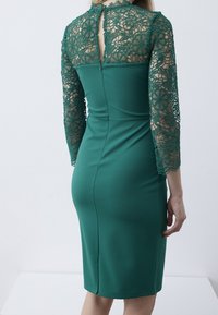Robe verte avec manches longues en dentelle délicate, une silhouette ajustée et une fermeture éclair au dos. Tissu lisse avec une coupe longueur genou.
