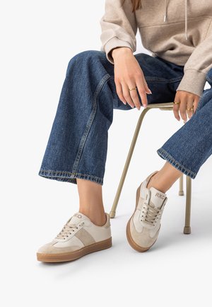 Persoană purtând adidași bej și albi cu talpă gumă, blugi albaștri și hanorac bej, stând pe un scaun cu mâinile sprijinite pe genunchi.