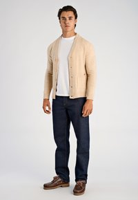 Beige stickad kofta med sjalkrage, knappstängning och framficka, kombinerad med mörkblå jeans och bruna snörskor.