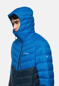 Giacca puffer blu con colletto alto e cappuccio, caratterizzata da una combinazione di tessuti lisci e trapuntati, con dettagli neri nella parte inferiore.