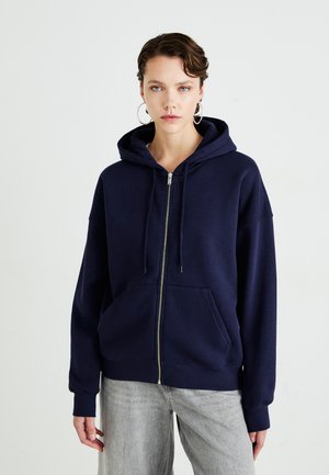 Giovane con i capelli corti che indossa grandi orecchini a cerchio, una felpa blu navy con zip e jeans grigio chiaro, su uno sfondo bianco.
